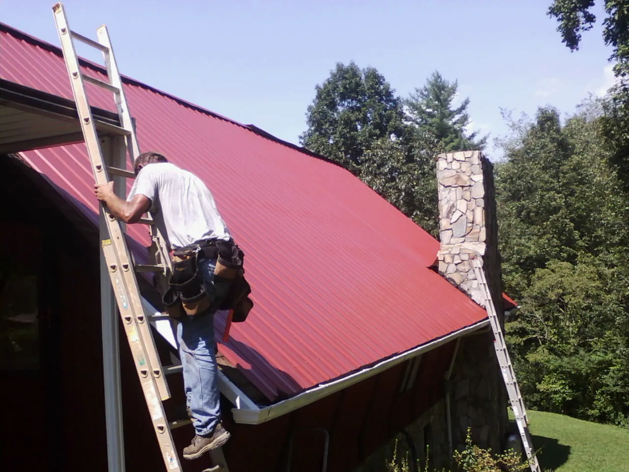 Roofer on a ladder beside a red metal roof and stone chimney — Soffit & Fascia Repair by Roofing Brothers in Tarzana